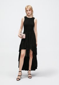 Robe noire à volants avec un corsage ajusté, une longueur asymétrique et un tissu texturé. Associée à un petit sac à main rose et des talons noirs à lanières.