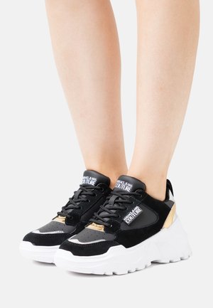 Baskets épaisses noir et blanc avec des accents dorés et le logo « Versace Jeans Couture » portées sur des jambes nues sur fond blanc.