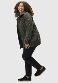 Sudadera verde con cremallera y textura estampada, con cordones en la capucha y acentos de logo. Combinada con jeans negros y zapatillas negras.