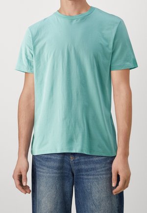 Hombre que lleva una camiseta de manga corta de color verde menta lisa y vaqueros azules contra un fondo neutro.