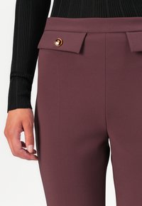 Calças cor burguês com uma textura suave, apresentando uma cintura dobrada e um detalhe com botão dourado, combinadas com um top de manga longa preta com nervuras.