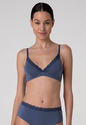 Giovane donna con capelli scuri che indossa un reggiseno blu con bordi in pizzo e mutandine a vita alta abbinate, in piedi davanti a uno sfondo chiaro e uniforme.
