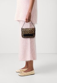 Henkilö vaaleanpunaisessa pörröisessä asusteessa pitää pientä leopardikuvioista käsilaukkua, jalassaan beiget ballerinat, seisten yksinkertaista valkoista taustaa vasten.