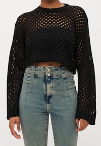 Maglione nero corto in maglia con motivo a trama aperta e maniche lunghe; abbinato a jeans a vita alta azzurri con effetto slavato.