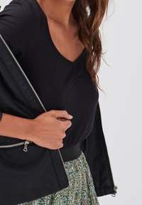 Haut noir à côtes avec un col en V, associé à une jupe verte à motifs et une veste noire zippée révélant des détails sur les côtés.