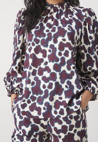 Blusa a maniche lunghe con scollo arricciato, di colore bianco con un motivo astratto multicolore in rosso e blu, con polsini elasticizzati.