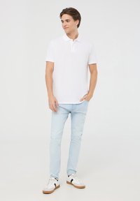 Polo bianca, maniche corte, con patta di chiusura a tre bottoni. Jeans slim azzurri. Sneakers bianche con dettagli neri. Sfondo neutro.