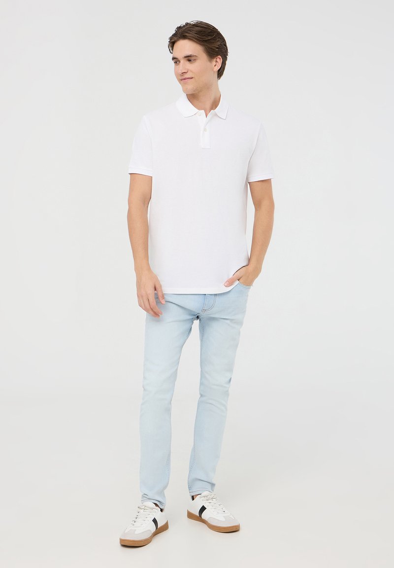 Polo bianca, maniche corte, con patta di chiusura a tre bottoni. Jeans slim azzurri. Sneakers bianche con dettagli neri. Sfondo neutro.