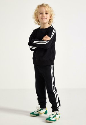 Sort sweatshirt og bukser med hvide striber på siden, kombineret med grønne og gule sneakers. Materialet virker blødt; pasformen er løs.