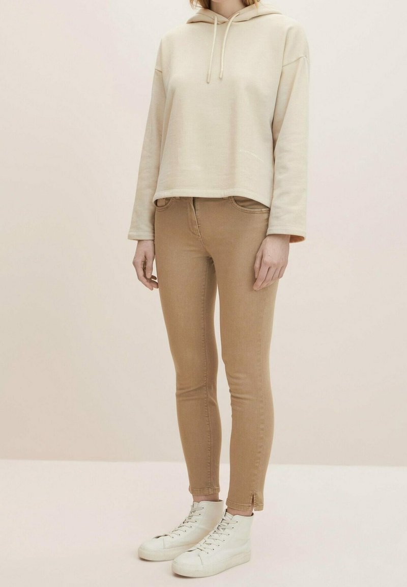 Sudadera corta beige con cordones, combinada con jeans ajustados marrón claro y zapatillas altas blancas. Texturas suaves y diseño ajustado.