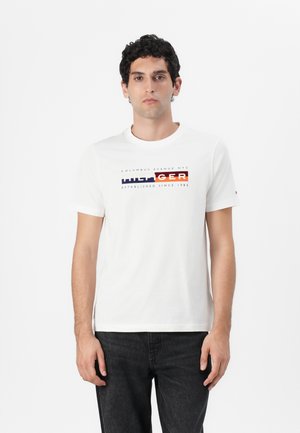 Bílá bavlněná tričko s krátkým rukávem, na kterém je barevný design loga "TOMMY HILFIGER" přes hrudník. Standardní kulatý výstřih.