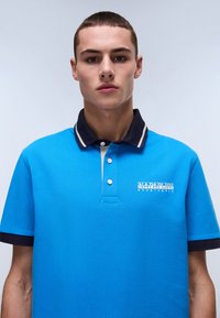 Blå polo skjorta i strukturerad tyg, med en marinblå krage med vita ränder, treknappsslutning och en logotyp på vänster bröst.