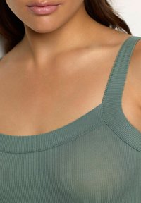 Ingrandimento di una persona che indossa una canotta a costine di un leggero verde, mostrando il scollo, le spalline e la parte superiore del petto.