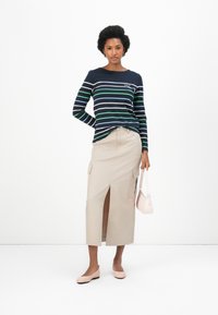 Marineblauwe en groene gestreepte blouse met lange mouwen, gecombineerd met een beige cargorechtersrok met een voorkijklits. Lichtroze flats en een handtas maken de look af.