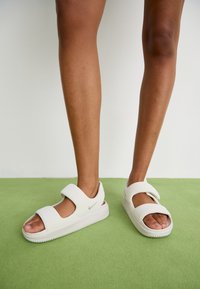 Valkoiset sandaalit, joissa on kaksi säädettävää nauhaa, tekstuuripohjat ja Nike-logo; valmistettu sileästä materiaalista ja avoimesta kärjestä.