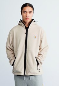 Chaqueta de forro polar beige con una cremallera negra, dos bolsillos laterales y una capucha ajustable. Presenta un pequeño logo en el área del pecho izquierdo.