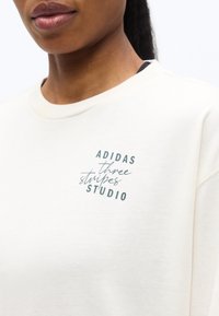 Κοντινό πλάνο ενός ατόμου που φοράει λευκό μπλουζάκι με στρογγυλή λαιμόκοψη και το κείμενο «ADIDAS three stripes STUDIO» τυπωμένο στο αριστερό μέρος του στήθους.