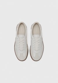 Baskets en suède blanc avec une semelle en caoutchouc brun, présentant un bout arrondi, un laçage sur le devant et un logo embossé sur la languette.