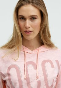 Hellrosa Kapuzenpullover mit Kordelzug, versehen mit einem großen Logo in einem dunkleren Farbton, akzentuiert durch Strasssteine zur Detailverfeinerung. Weiche Textur.