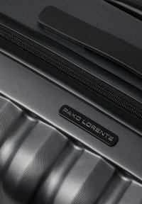 Zbliżenie na teksturowaną ciemnoszarość walizkę z czarnym zamkiem błyskawicznym, wyściełanym uchwytem i prostokątną metką marki "PAKO LORENTE".