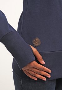 Högra handen vilar på höften, iförd en marinblå sweatshirt med en liten triangulär patch-logotyp och mörka jeans.