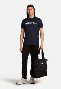Marine T-shirt met witte bergafbeelding, gecombineerd met zwarte joggers, en een zwarte nylon draagtas in de hand. Witte sneakers met grijze accenten.