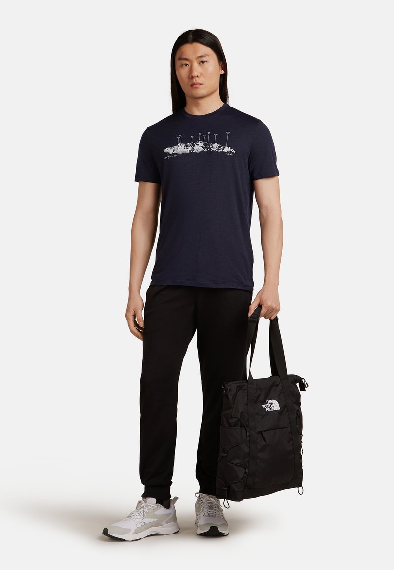 Marine T-shirt met witte bergafbeelding, gecombineerd met zwarte joggers, en een zwarte nylon draagtas in de hand. Witte sneakers met grijze accenten.