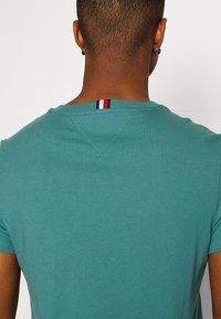 Person med kort hår og små hoopsøreringe iført en teal t-shirt med rød, hvid og blå stofstribe ved nakkeåbningen.