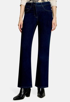 Person iført mørkeblå vidde jeans kombineret med sorte ankelstøvler med hæl, stående mod en ensartet lys baggrund.