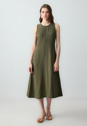 Sleeveless olive green dress made of linen, featuring a fitted bodice, flared skirt, and subtle seam detailing. Worn with flat sandals.