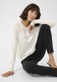 Helles cremefarbenes Langarmpullover mit rundem Hals und gerippten Bündchen, kombiniert mit schwarzen, figurbetonten Jeans und offenen Absatzschuhen, in sitzender Pose.