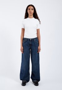 Vitt kortärmat T-shirt som matchas med vida blå jeans. T-shirten har en ribbad rund hals; jeansen har framfickor och en knappstängning.