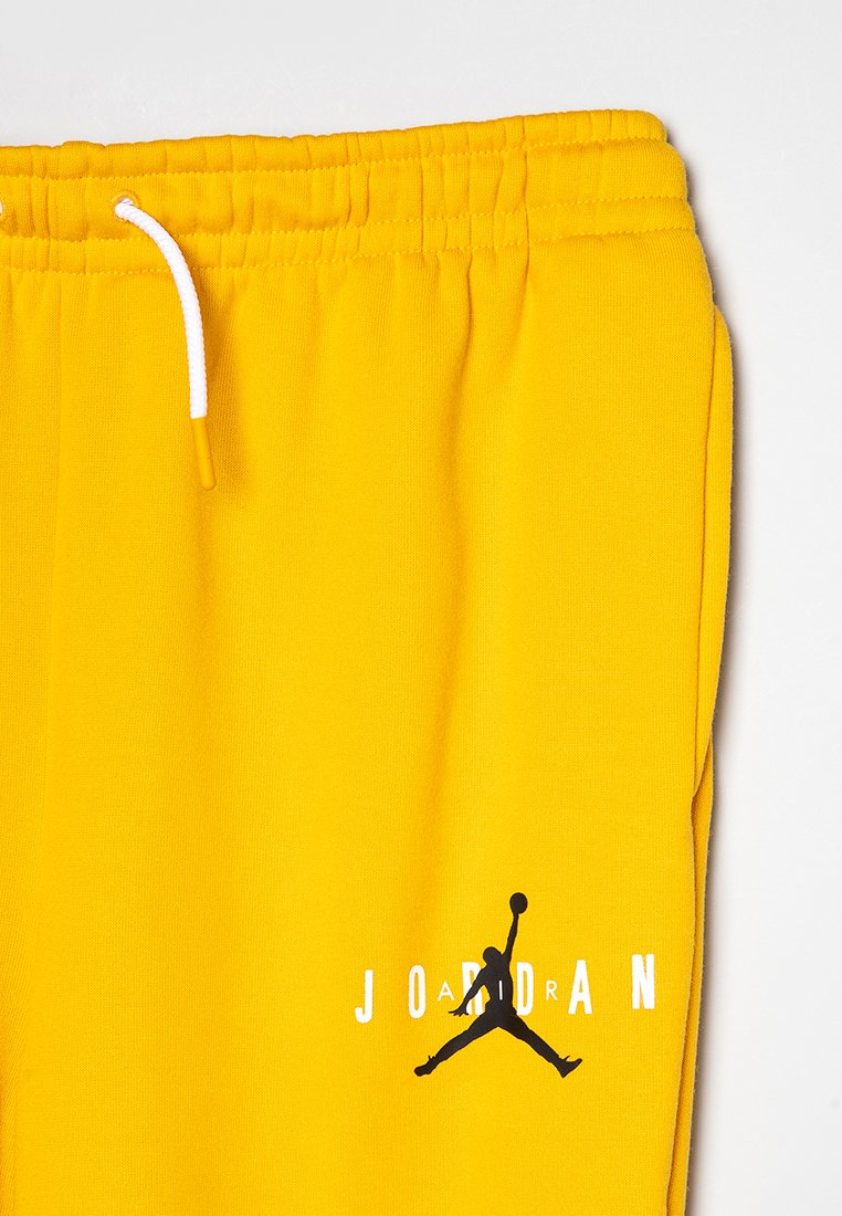 Gelbe Jogginghosen aus weichem Material. Sie verfügen über einen elastischen Bund mit weißen Kordeln und einem schwarzen Logo mit dem Text "JORDAN".
