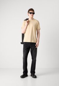 Beige bomuld t-shirt med logo, sorte denim jeans og sorte slip-on sko. Modellen har mørke solbriller på og holder en sort jakke.