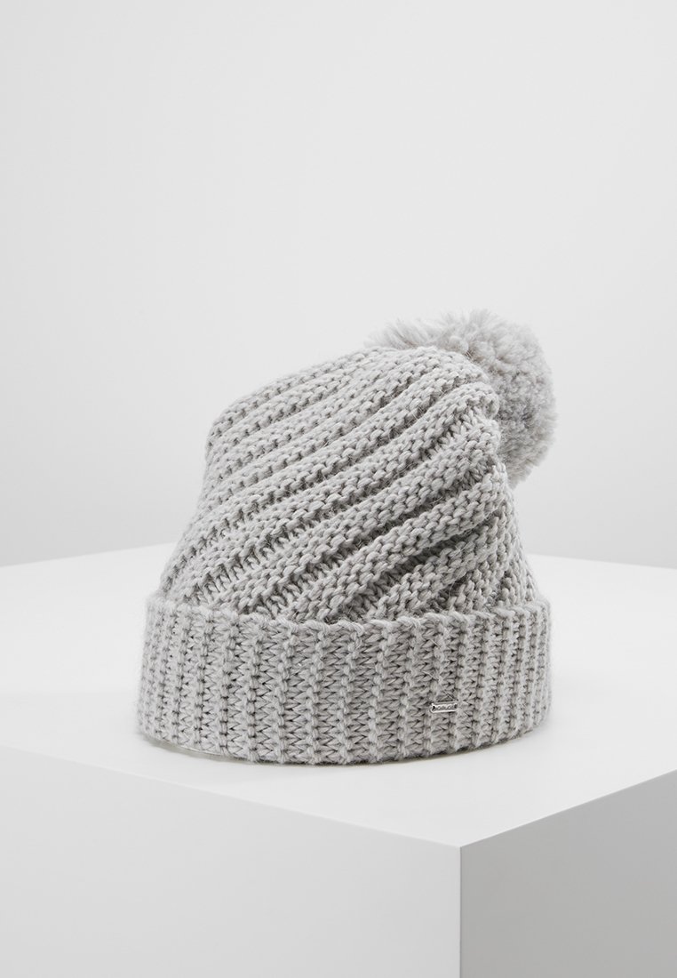 Bonnet d'hiver gris tricoté avec bord-côtes et pompon sur le dessus, posé sur une surface blanche sur fond gris clair.