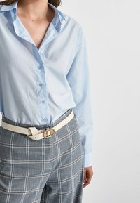 Femme portant une chemise bleu clair boutonnée rentrée dans un pantalon gris à carreaux avec une ceinture blanche avec une boucle double anneau dorée.