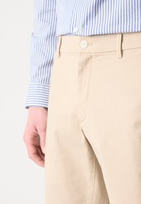 Beige bomullsbukser har en jevn tekstur, frontknappelukking og sidelommer. En blå og hvit stripete skjorte er delvis synlig.