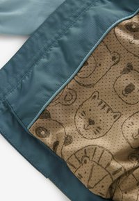 Intérieur d'une veste bleu clair avec un tissu beige arborant un motif de visages d'animaux et de points. Texture lisse, bordure bleu contrastante.