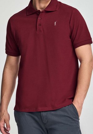 Mężczyzna w bordowej koszulce polo z krótkim rękawem, z małym wyszywanym logo golfisty na piersi oraz ciemnoszarych spodniach.