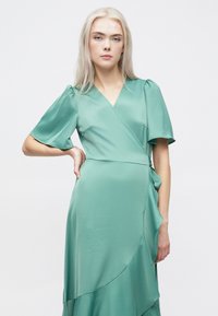 Πράσινο σατέν φόρεμα με φουσκωτά μανίκια, ασύμμετρο στρίφωμα και μαλακή ραφή. Διαθέτει v-λαιμόκοψη και λεπτομέρεια με ζώνη στο πλάι.