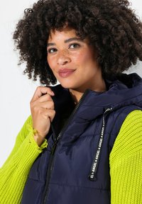 Marinblå puffväst med en strukturerad huva, zip-förslutning och dragsnören. Kombinerad med en ljusgrön stickad tröja med lös väv och fransade ärmslut.