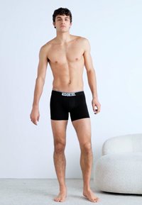 Boxer-briefs noirs de Diesel avec une ceinture élastique estampillée. Le modèle se tient pieds nus contre un fond clair.