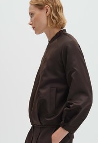 Vrouw met kort blond haar, gekleed in een donkerbruine jas met rits en bijpassende broek, staande in profiel met handen in de zakken, neutrale achtergrond.