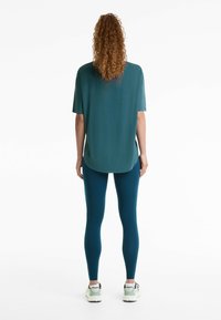 Haut turquoise à manches courtes avec fentes latérales, associé à des leggings turquoise foncé. La tenue est complétée par des chaussures de sport de couleur claire.