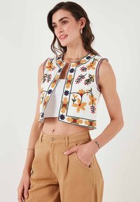 Cropped vest in white fabric with floral embroidery in red, blue, and yellow. Features a round neckline and open front design.
