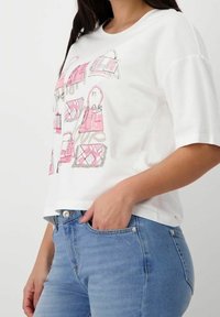 Bílá krátká tričko s krátkým rukávem s grafikou růžových kabelků. Má volný střih a je kombinováno se světle modrými džíny.
