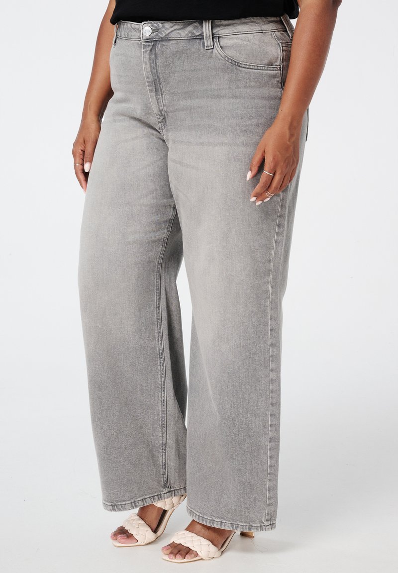 Jean gris à jambes larges en denim, à taille haute et avec un design à cinq poches. Porté avec des sandales tissées beiges.