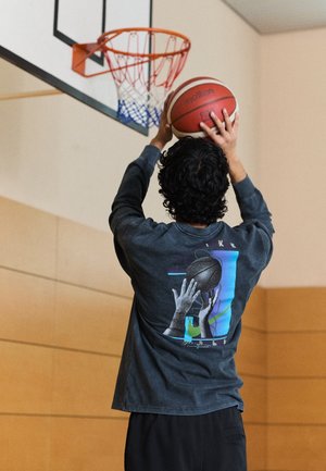 Persoon die een donker shirt met lange mouwen draagt, bereidt zich voor om een basketbal richting een binnenspeelkorf in een sporthal te schieten.