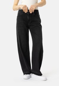 Osoba v širokých černých džínách s t�řepenými okraji a bílými teniskami si zapíná džíny v pase.