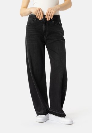 Person som har på seg vide svarte jeans med frynsete nederkant og hvite sneakers, og fester jeansene i midjen.